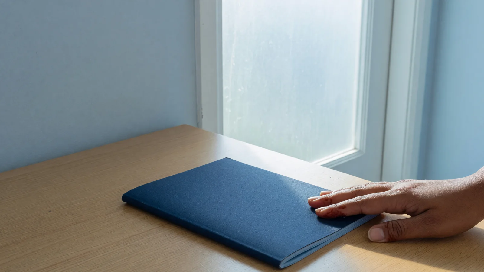 Adult hand on a closed booklet at a bright doorway desk